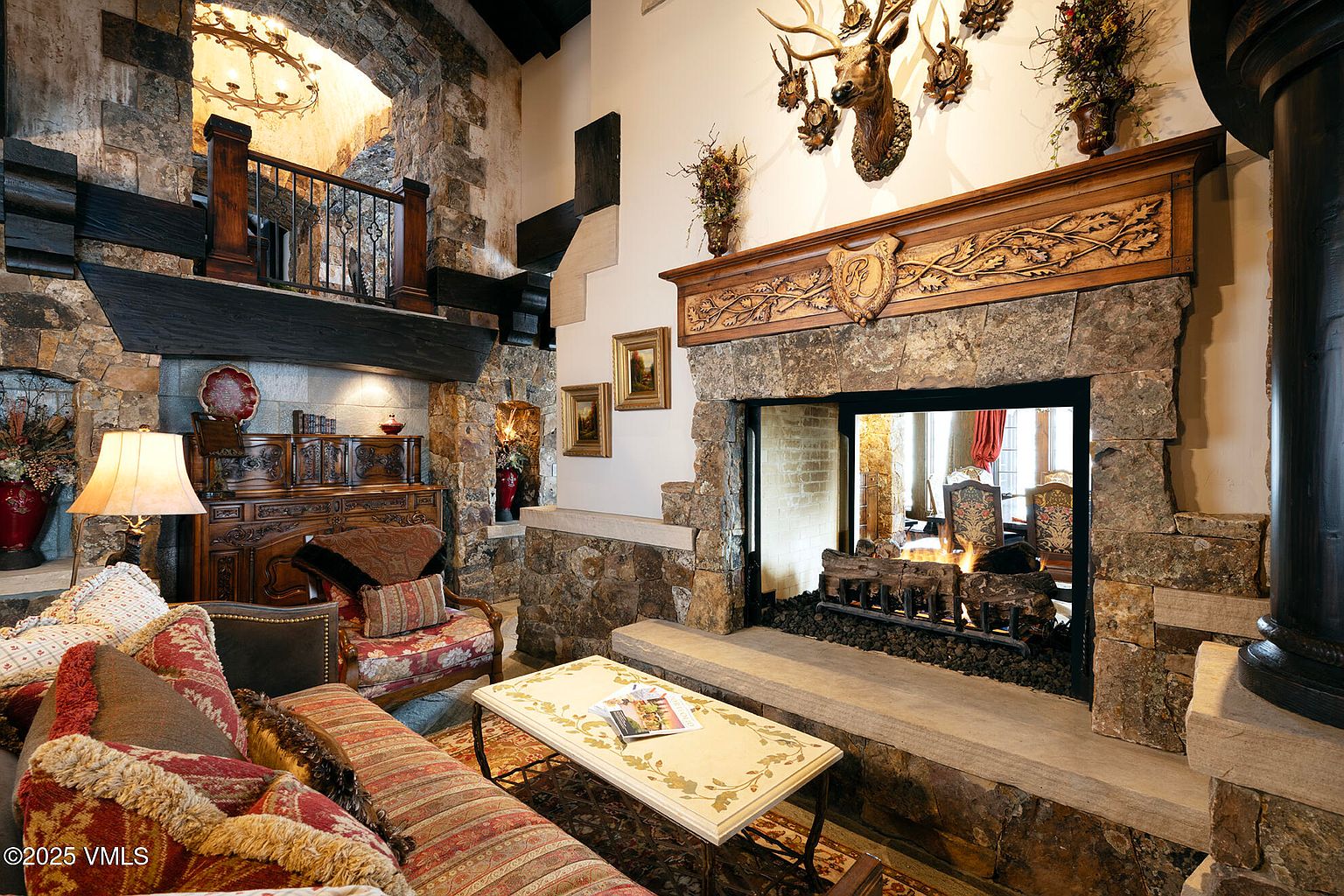 This rustic living room features a prominent stone-clad chimney breast with an ornate carved wood mantel and a mounted deer head above. The space is furnished with patterned upholstered seating, a light-colored coffee table, and a dark wood sideboard, all set against a backdrop of stone walls and exposed timber beams. A mezzanine level with a wrought-iron railing overlooks the area, contributing to the grand, lodge-inspired atmosphere.