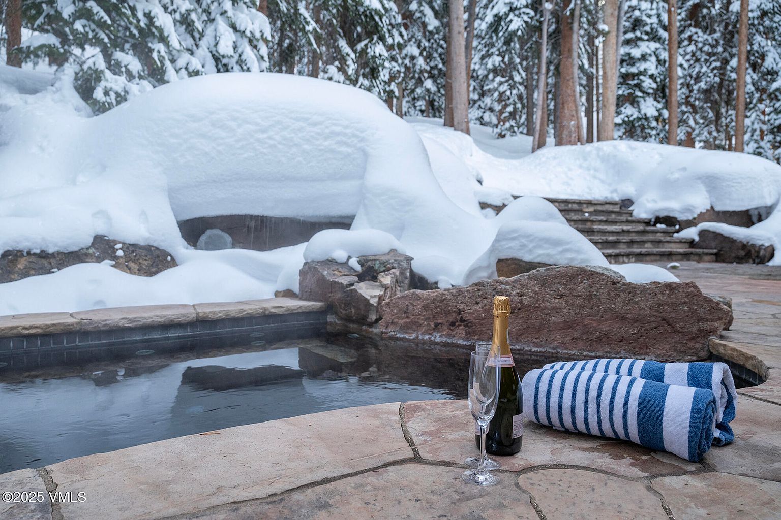 This image captures a serene outdoor spa area nestled in a snowy, wooded landscape. A stone-lined hot tub filled with steaming water sits in the foreground, accompanied by a bottle of champagne, two flutes, and a rolled blue-and-white striped towel on the stone patio. The background features deep snow banks and a set of stone steps leading up into the forest, creating a luxurious and tranquil winter retreat atmosphere.