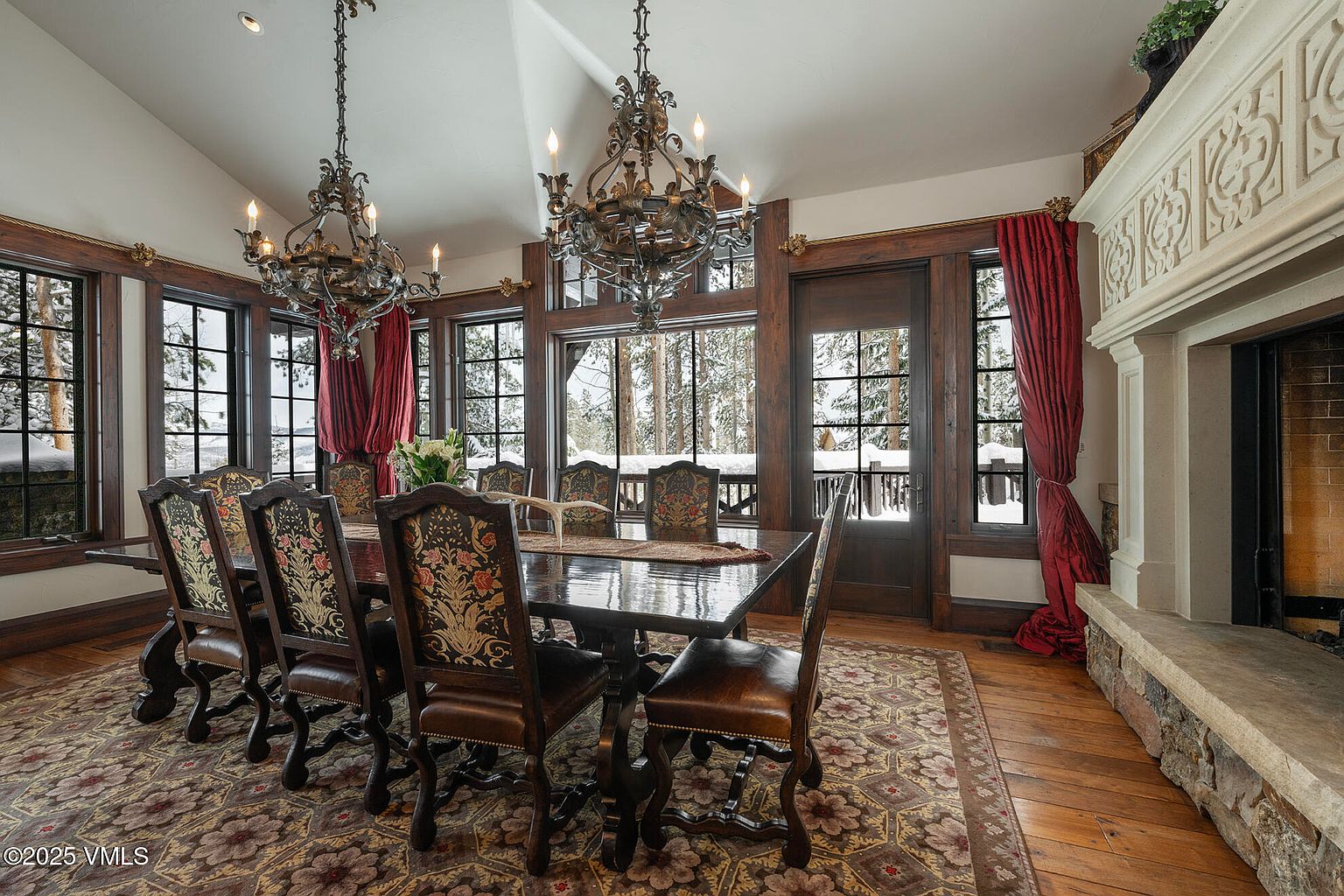This elegant dining room features a large, dark wood table surrounded by ornate chairs with floral-patterned upholstery. The space is illuminated by two elaborate, multi-tiered iron chandeliers and framed by tall windows that offer views of a snowy, wooded landscape. A stone-clad fireplace adds warmth to the room, which is finished with rich wood trim, deep red curtains, and a detailed patterned area rug.