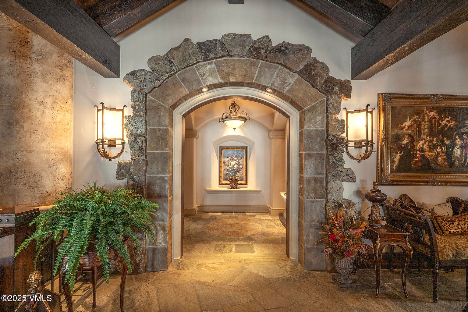 This elegant hallway features a prominent stone-clad archway that serves as a dramatic transition between living spaces. The area is adorned with ornate wall sconces, a framed painting, and a decorative ceiling light, all set against a backdrop of stone flooring and rustic exposed wood beams. The perspective is centered, drawing the eye through the stone arch into a secondary gallery space, creating a sophisticated and Old World atmosphere.