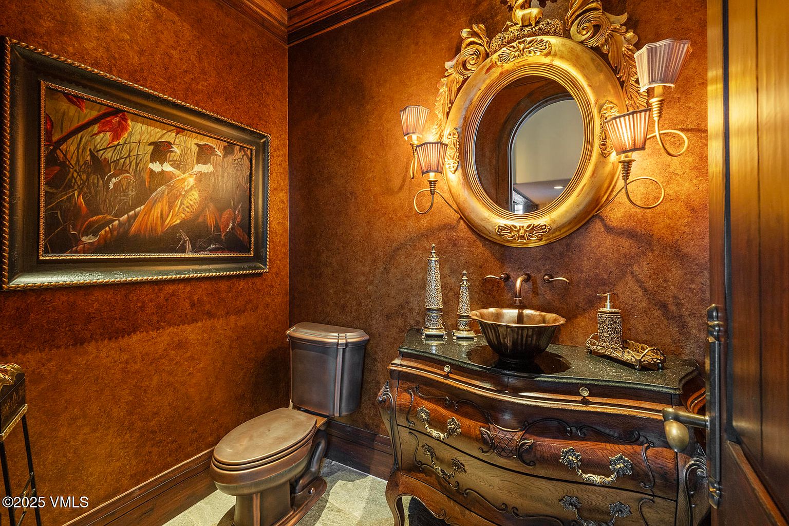 This ornate powder room features rich, textured brown walls and a sophisticated, antique-style vanity with a vessel sink. A large, gilded circular mirror serves as the focal point, flanked by elegant wall sconces, while a framed wildlife painting adds a touch of traditional character. The space is completed by a dark-toned toilet and a stone-topped vanity, creating a moody and luxurious atmosphere.