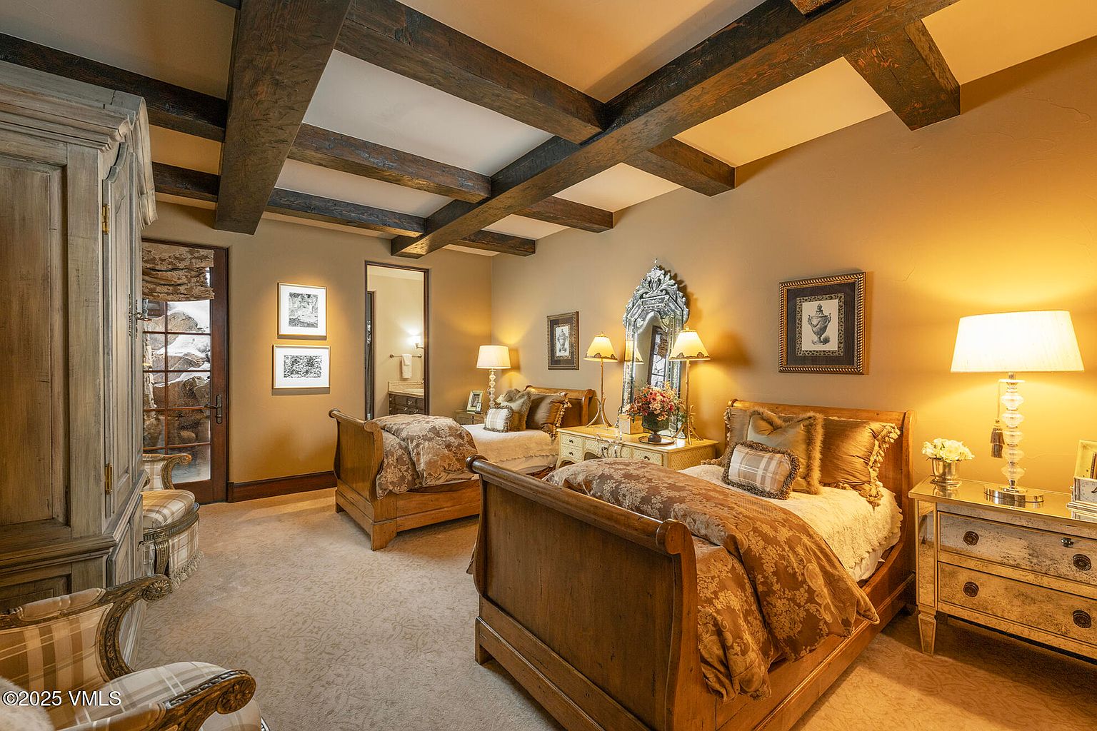 This elegant guest bedroom features two wooden sleigh beds with patterned bedding, set beneath a striking ceiling with exposed dark wood beams. The room is warmly lit by multiple table lamps and includes a decorative mirror, framed artwork, and a nightstand with a stone top. A doorway leads to an adjacent bathroom, and the overall aesthetic is traditional and cozy with a neutral color palette.