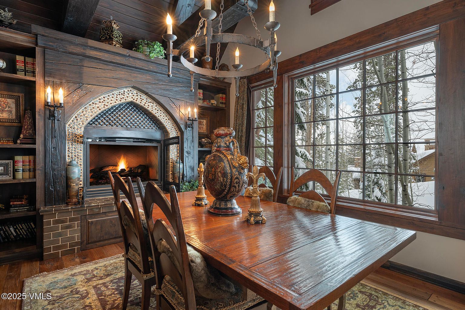 This rustic dining room features a heavy wooden table surrounded by chairs with fur-lined seats, set beneath an ornate iron chandelier. A prominent fireplace with a decorative tiled arch serves as the focal point, flanked by built-in wooden shelving filled with books. Large windows provide a scenic view of a snow-covered forest, creating a cozy and sophisticated mountain lodge atmosphere.