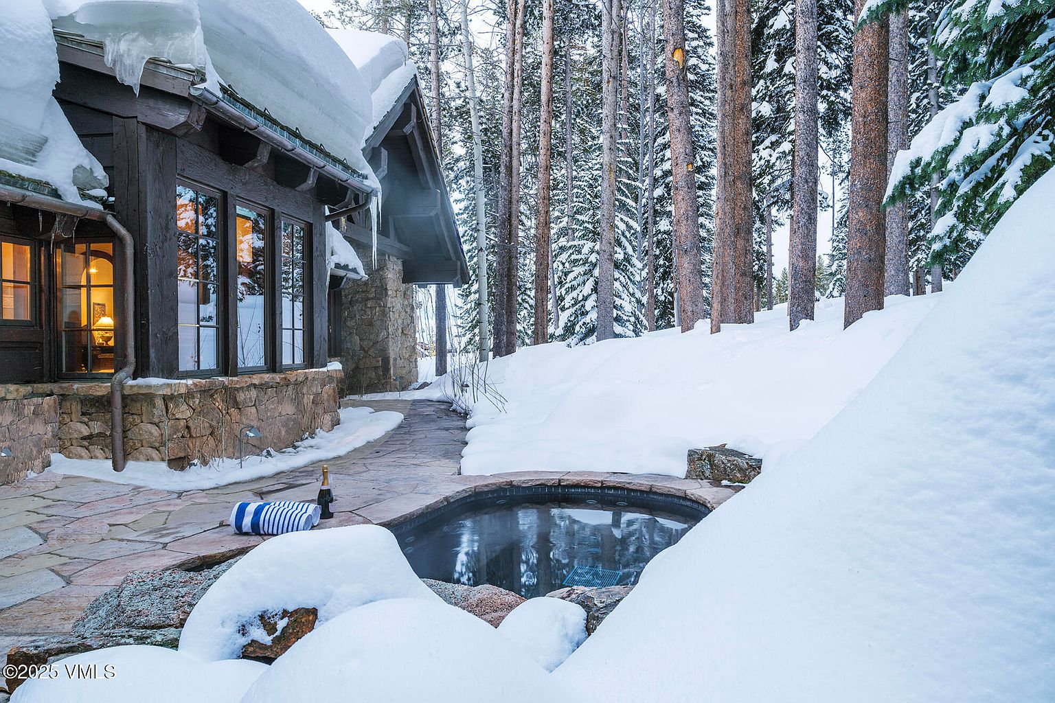 This exterior view features a stone-clad spa nestled into a snow-covered patio area adjacent to a rustic mountain home. The structure showcases dark wood siding and large windows that reflect the surrounding pine forest, creating a cozy, secluded atmosphere. A bottle of champagne and rolled towels sit on the stone patio, emphasizing a luxurious, relaxing retreat experience.