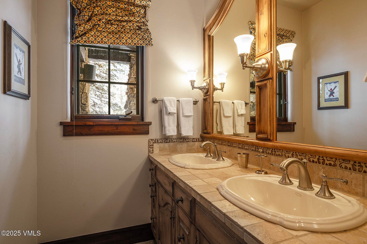 This rustic-style bathroom features a dual-sink vanity with a stone-tiled countertop and dark wood cabinetry. A large, wood-framed mirror reflects the space, which is accented by patterned window treatments and framed ski-themed artwork on the walls. The warm lighting and natural textures create a cozy, mountain-lodge aesthetic.