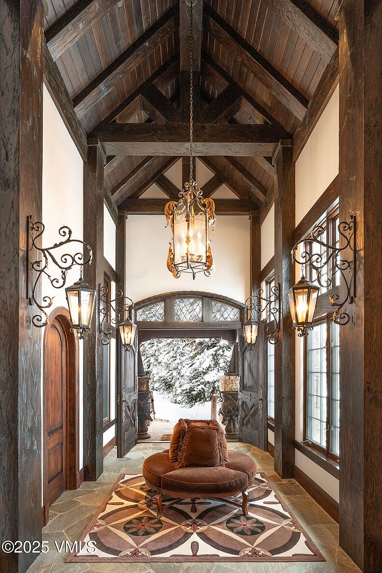 This grand entryway features a dramatic vaulted ceiling with exposed dark wood beams and a central ornate lantern chandelier. The space is defined by rich wood trim, stone flooring, and symmetrical wall-mounted lanterns that lead the eye toward an arched doorway revealing a snowy exterior. A circular velvet ottoman sits atop a decorative patterned rug, creating a sophisticated and welcoming focal point in this rustic, mountain-inspired foyer.