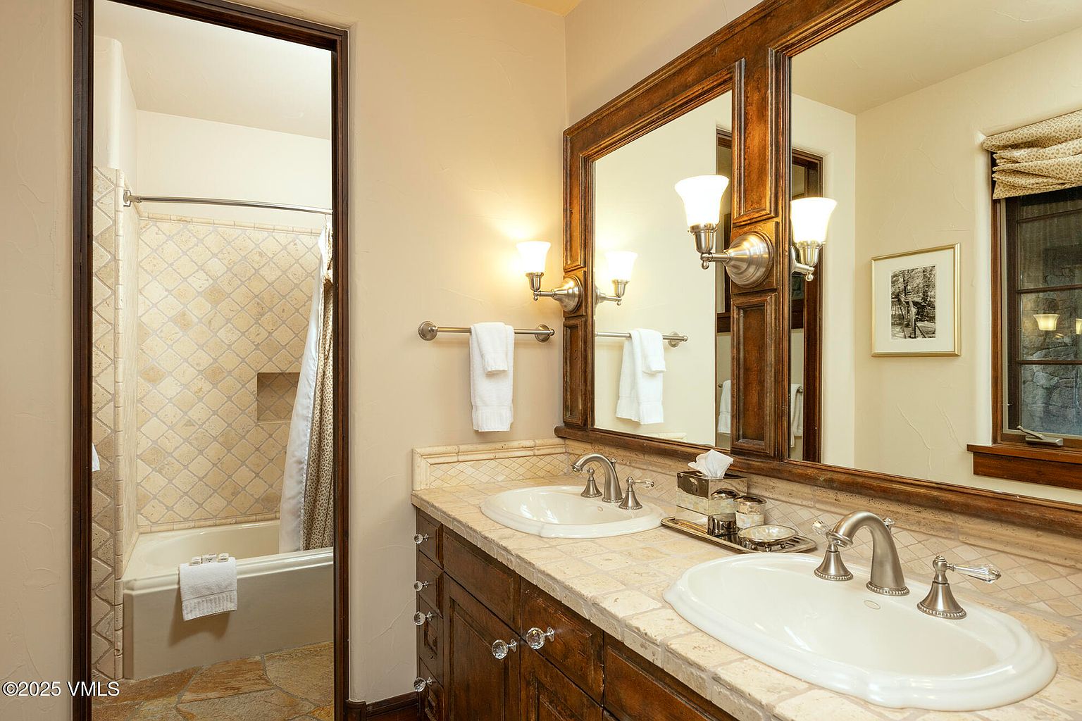This bathroom features a dual-sink vanity with a stone countertop and dark wood cabinetry, complemented by a large, framed mirror and wall-mounted light fixtures. To the left, a full-length mirror reflects a tiled shower and tub combination with a neutral-toned shower curtain. The space is finished with light-colored wall paint and stone tile flooring, creating a warm and traditional aesthetic.