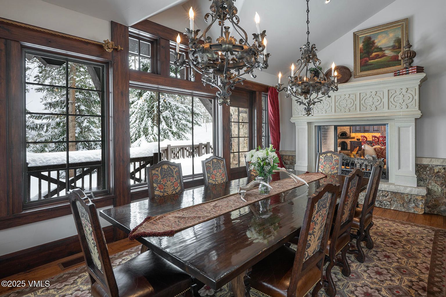 This elegant dining room features a long, polished dark wood table surrounded by high-backed chairs with ornate patterned upholstery. The space is anchored by two large, intricate iron chandeliers and a prominent stone fireplace with a detailed carved surround. Large windows along the side wall offer a scenic view of a snow-covered landscape, creating a cozy and sophisticated atmosphere.