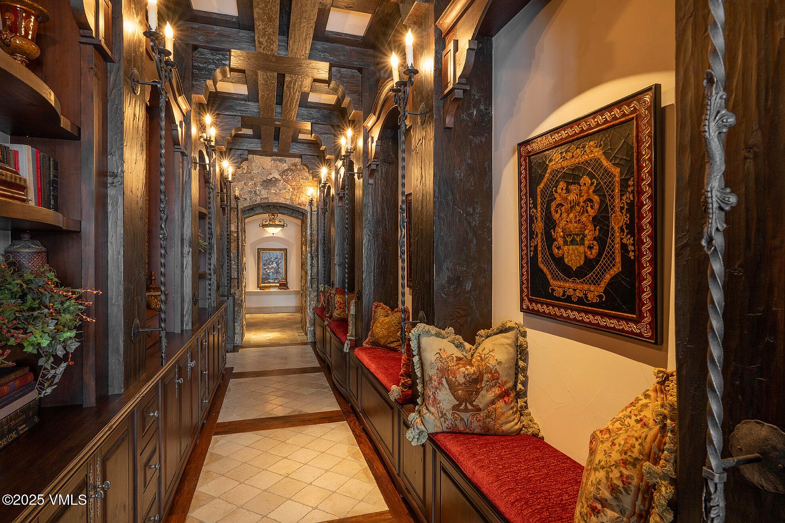 This ornate, castle-inspired hallway features dark, heavy wood paneling, exposed ceiling beams, and a series of wall-mounted iron sconces. A built-in bench with plush red velvet cushions runs along the right wall, accented by decorative pillows and a large, framed crest hanging above. The perspective looks down the length of the hall, past a stone-arched transition, toward a brightly lit alcove containing a piece of artwork.