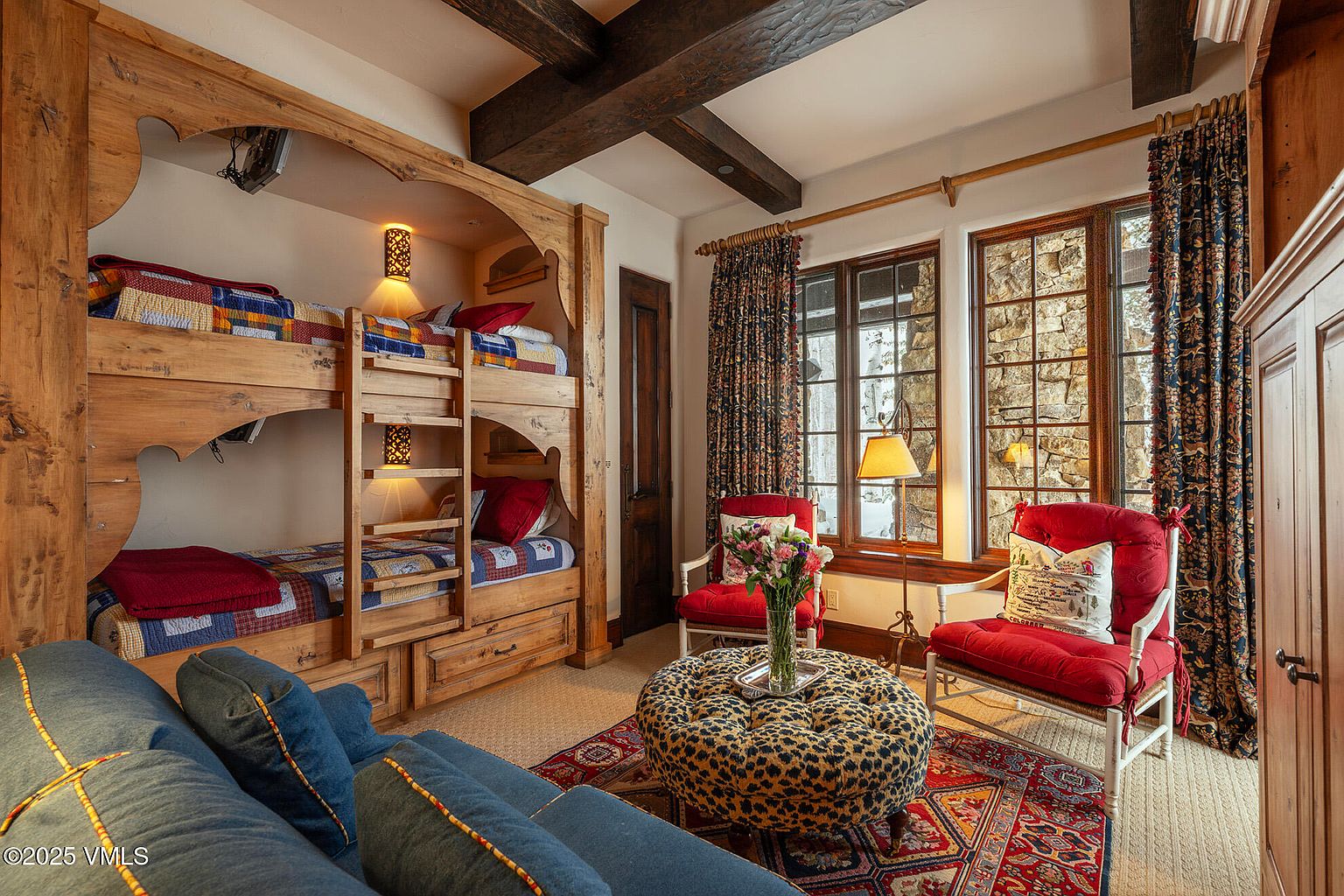 This cozy bedroom features a custom-built wooden bunk bed unit with integrated lighting and a rustic aesthetic. The room is furnished with a blue sofa, two red armchairs, and a leopard-print ottoman centered on a vibrant patterned rug. Large windows with dark patterned curtains allow natural light to illuminate the space, which is accented by exposed wooden ceiling beams.
