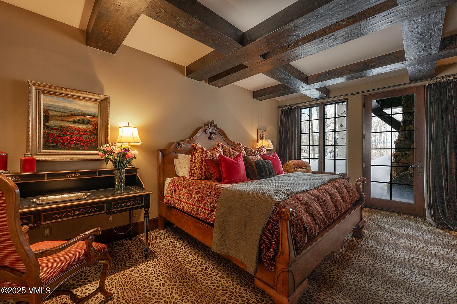 This cozy bedroom features a prominent wooden bed frame with ornate detailing, dressed in rich red and patterned textiles. The room is accented by dark exposed ceiling beams, a leopard-print carpet, and a classic writing desk topped with a floral arrangement and a framed landscape painting. A glass-paned door leads to an outdoor balcony, allowing natural light to filter into the warm, traditionally styled space.