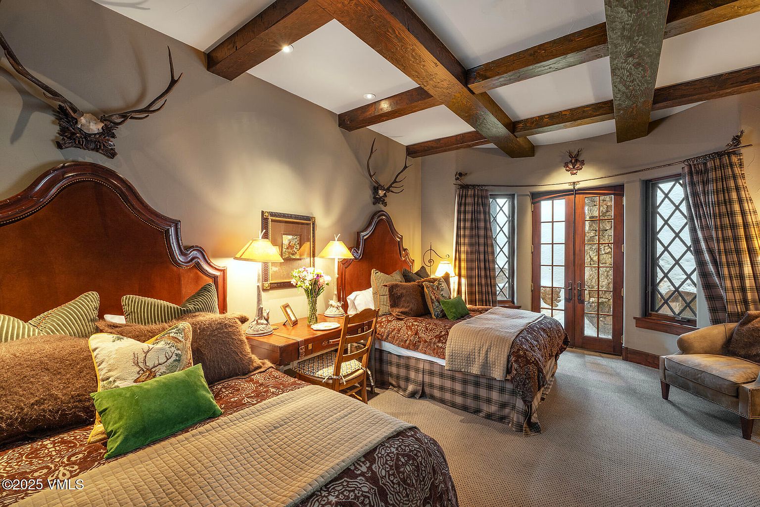 This rustic-style guest bedroom features two beds with ornate, dark wood headboards and patterned bedding. A shared wooden desk sits between the beds, illuminated by matching table lamps, while exposed wooden ceiling beams add architectural character to the space. Large glass-paned doors provide access to the outdoors, flanked by plaid curtains and decorative wall-mounted antlers.