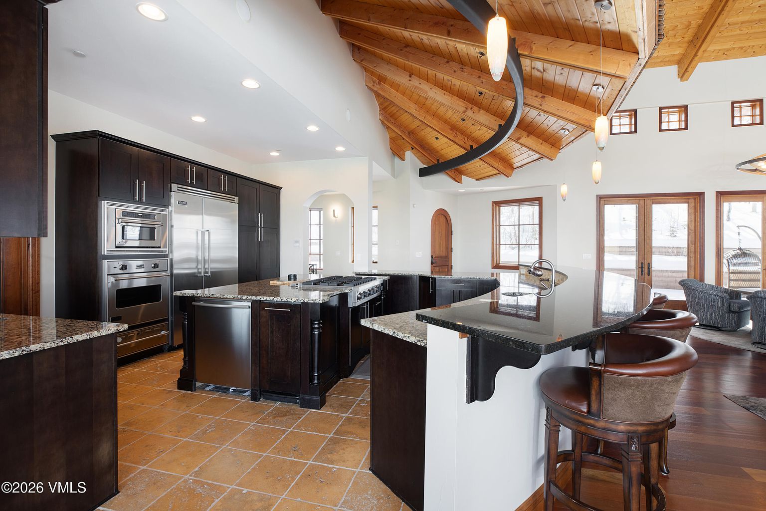 This is a spacious kitchen featuring dark wood cabinetry, stainless steel appliances, and granite countertops. A large island with a cooktop is centrally located, and a breakfast bar with seating is adjacent to a set of doors leading to an outdoor area. The ceiling is wood paneled with unique lighting fixtures, and the floor is tiled.