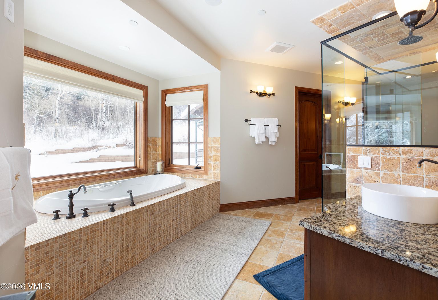 This is a luxurious primary bathroom featuring a large soaking tub situated beneath a wide window offering a view of a snowy landscape. The bathroom includes a vanity with a granite countertop and a vessel sink, along with a glass-enclosed shower. The overall design incorporates warm tones and natural materials, creating a spa-like atmosphere.