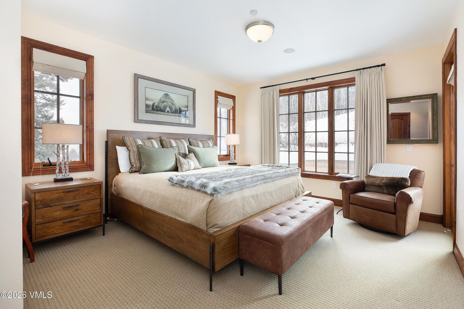 This is a primary bedroom featuring a large bed with a wooden frame, several pillows, and a faux fur throw. The room has two windows with wooden frames and white curtains, allowing natural light to fill the space. A leather bench sits at the foot of the bed, and a comfortable armchair is positioned near one of the windows, creating a cozy and inviting atmosphere.