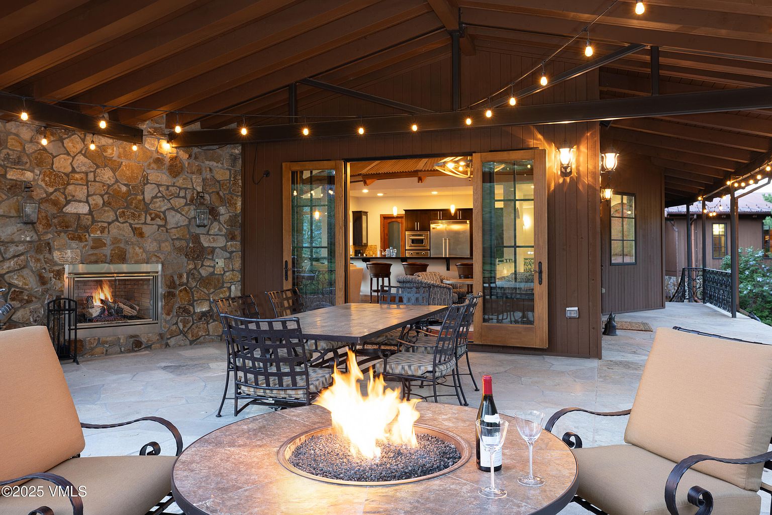This image showcases a beautifully designed outdoor patio area, perfect for entertaining or relaxing. A stone fireplace anchors one side, while a dining table and chairs offer a space for meals. In the foreground, a fire pit with a bottle of wine and glasses creates a cozy ambiance, enhanced by string lights overhead. The open doorway leads into the home, seamlessly blending indoor and outdoor living.