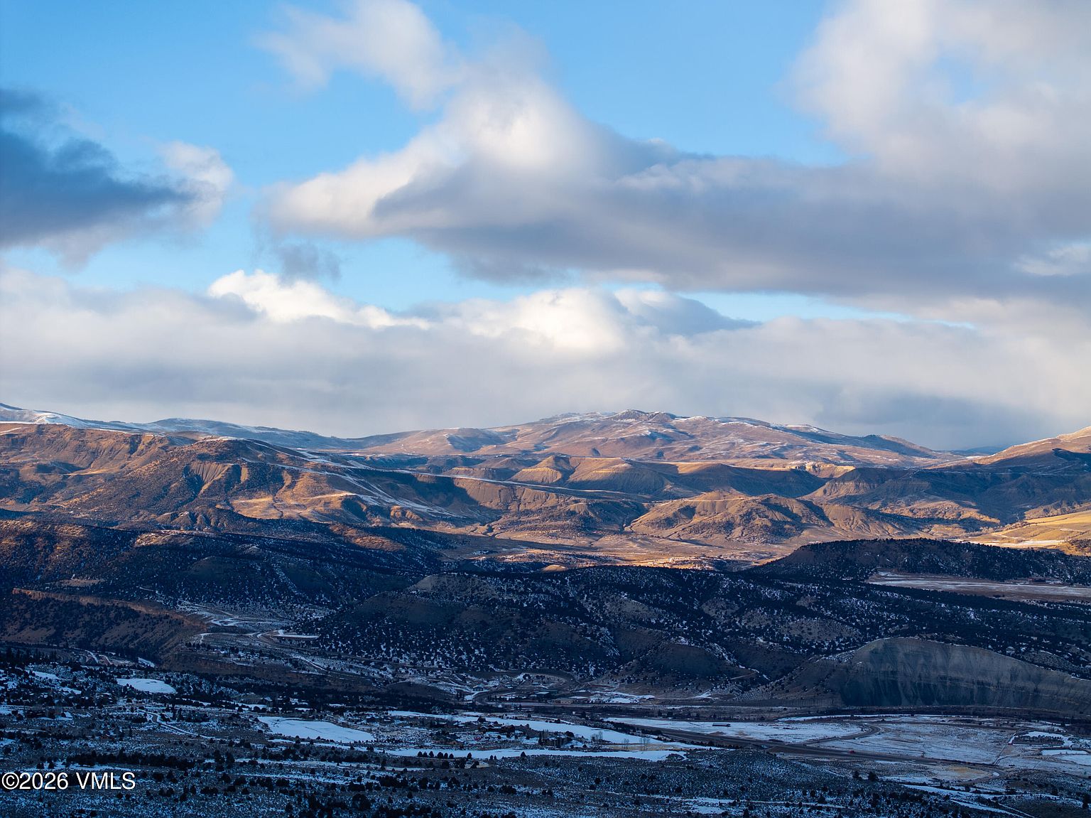 This aerial view showcases a vast landscape of rolling hills and valleys, partially covered in snow, under a bright blue sky with scattered clouds. The terrain is rugged and varied, with visible shadows accentuating the depth and texture of the land. This image provides a sense of the property's expansive surroundings and natural beauty.
