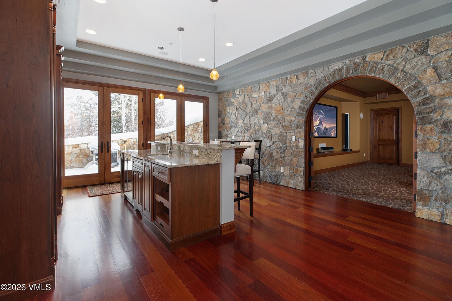 This interior shot showcases a kitchen area with rich hardwood floors and a central island featuring granite countertops. The kitchen seamlessly transitions into a stone-walled archway leading to another room, creating an open and inviting space. Natural light floods the area through large glass doors, offering a glimpse of the snowy exterior.