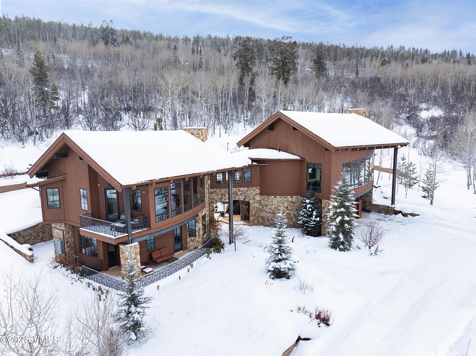 This aerial view showcases a luxurious mountain home with a brown exterior, stone accents, and snow-covered roof. The property is surrounded by a winter landscape with trees and a blanket of snow, creating a serene and secluded atmosphere. The architectural design features multiple levels and balconies, enhancing the home's appeal.