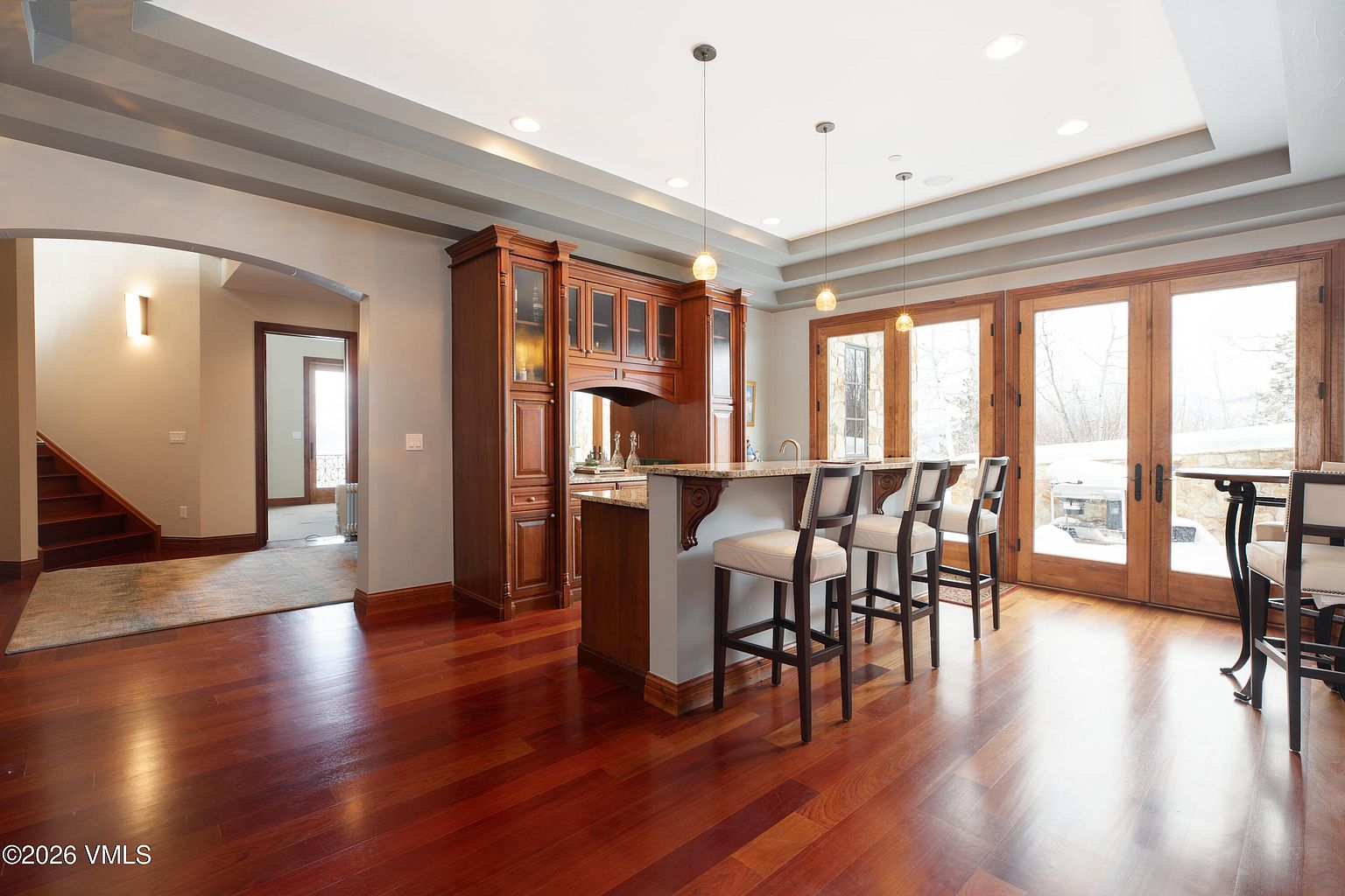 This interior shot showcases a well-appointed kitchen with rich wood cabinetry and a breakfast bar featuring bar stools. Natural light floods the space through large glass doors, highlighting the hardwood flooring. The kitchen seamlessly blends functionality with an elegant design, creating a warm and inviting atmosphere.