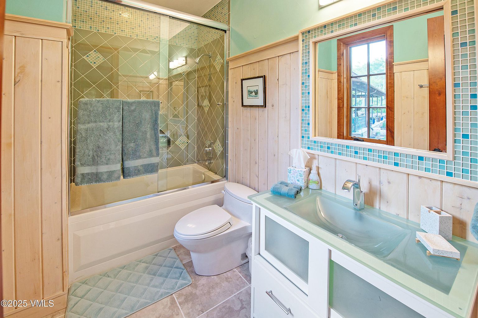 This is a well-lit bathroom featuring a bathtub with a glass enclosure, a toilet, and a vanity with a glass countertop. The walls are adorned with light-colored wood paneling and decorative tile accents. The overall style is clean and functional, with a touch of rustic charm.
