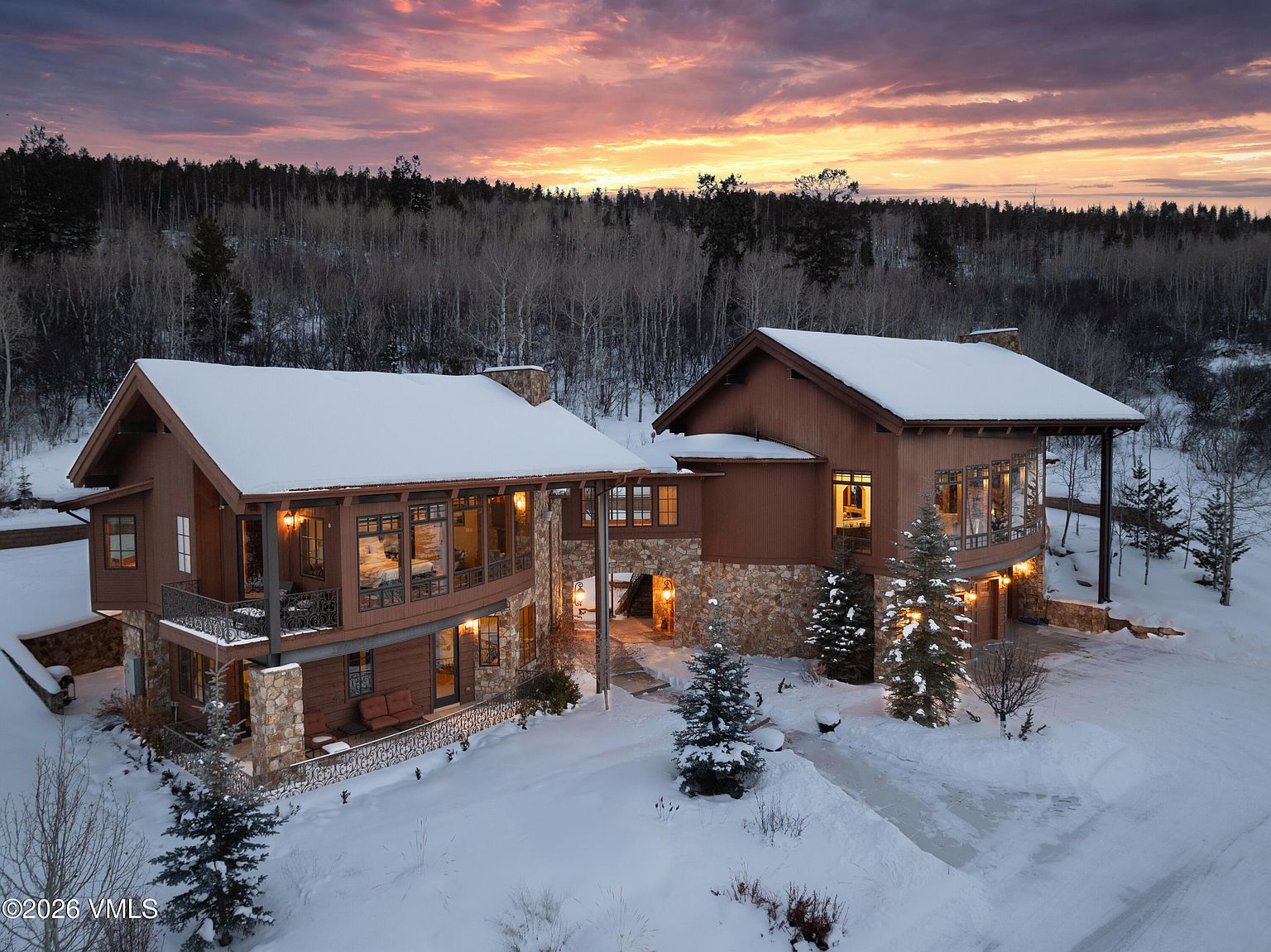 This aerial view showcases a luxurious mountain home in winter, featuring a stone and wood exterior with snow-covered roofs. The property is surrounded by a forest backdrop under a vibrant sunset sky, creating a serene and picturesque setting. The driveway is partially cleared, leading to the entrance of the home.