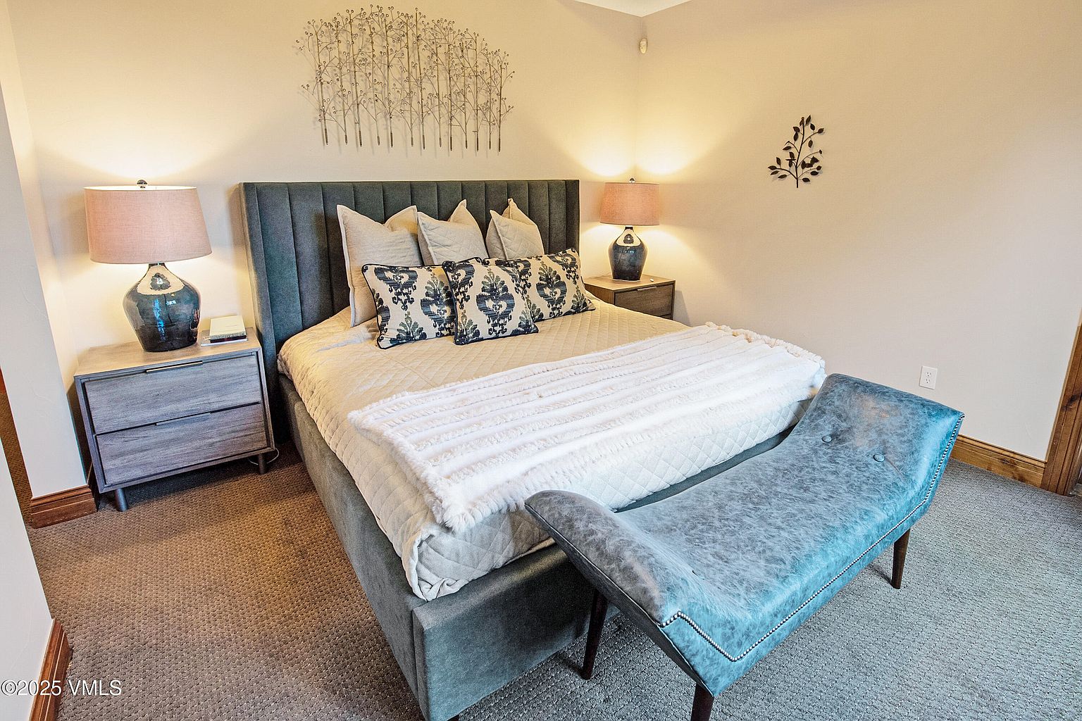 This is a cozy primary bedroom featuring a large bed with a tufted headboard, several decorative pillows, and a soft throw blanket. Two bedside tables with lamps flank the bed, and a bench sits at the foot of the bed. The room is carpeted and decorated with wall art, creating a warm and inviting atmosphere.