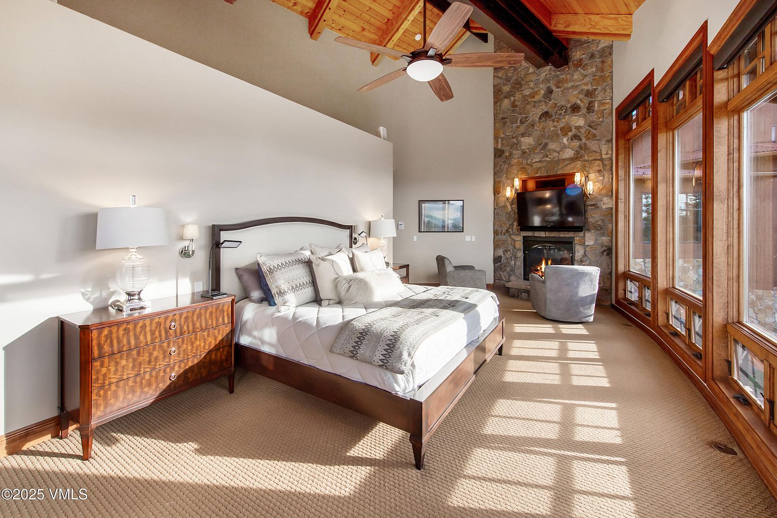 This is a luxurious primary bedroom featuring a king-size bed with a padded headboard, a stone fireplace with a mounted television, and a wall of windows providing ample natural light. The room is decorated in neutral tones with wood accents, creating a warm and inviting atmosphere. The perspective is from the doorway, showcasing the entire room.