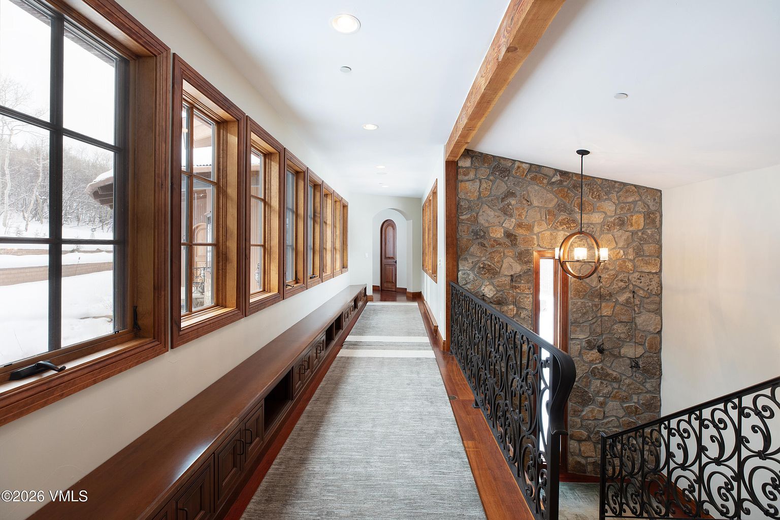 This interior shot showcases a well-lit hallway with a series of windows providing natural light. A long, built-in bench runs along the wall beneath the windows, offering seating and storage. The hallway leads to a staircase with ornate iron railings, and a stone accent wall adds a touch of rustic elegance to the space.