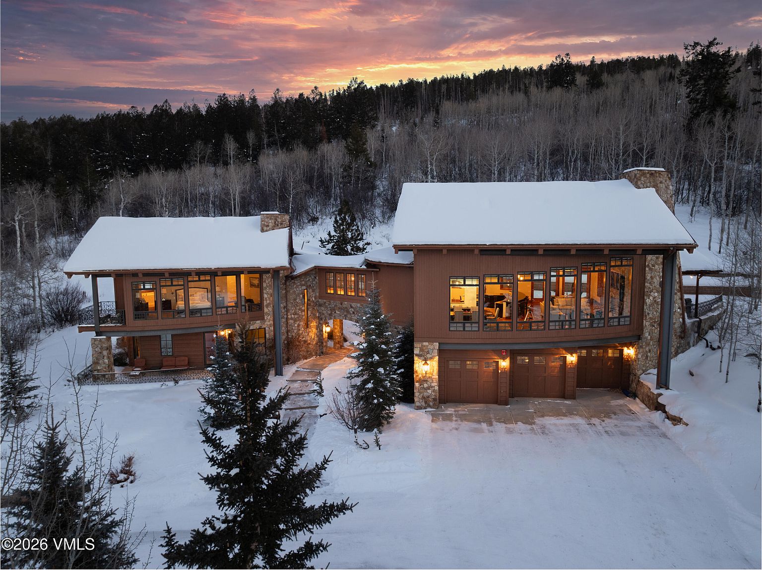 This is an eye-level, cinematic shot of a luxurious mountain home in winter, showcasing its grand facade with a stone and wood exterior. The house features multiple levels, large windows offering interior views, and a snow-covered roof, complemented by the surrounding snow-covered landscape and evergreen trees. The warm glow from the interior lights creates an inviting atmosphere against the backdrop of a colorful sunset sky.