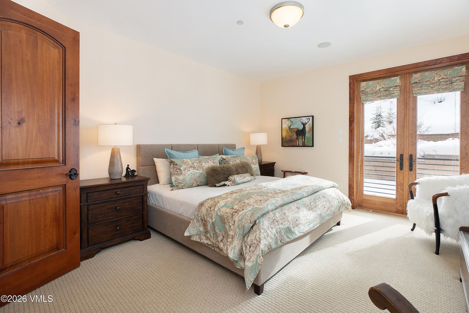 This is a cozy guest bedroom featuring a queen-sized bed with a patterned duvet and throw pillows. The room has neutral-colored walls and carpet, with a wooden door and a set of French doors leading to a balcony covered in snow. The room is well-lit with a ceiling light and two bedside lamps, creating a warm and inviting atmosphere.