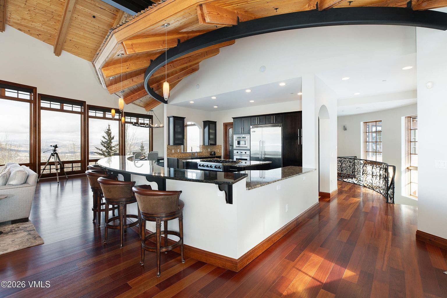This is an interior shot of a luxurious kitchen featuring dark wood cabinetry and a large, curved island with bar seating. The kitchen is open to a living area with large windows offering scenic views. A unique architectural detail is the curved wooden ceiling with a black metal support beam, adding a modern touch to the space.