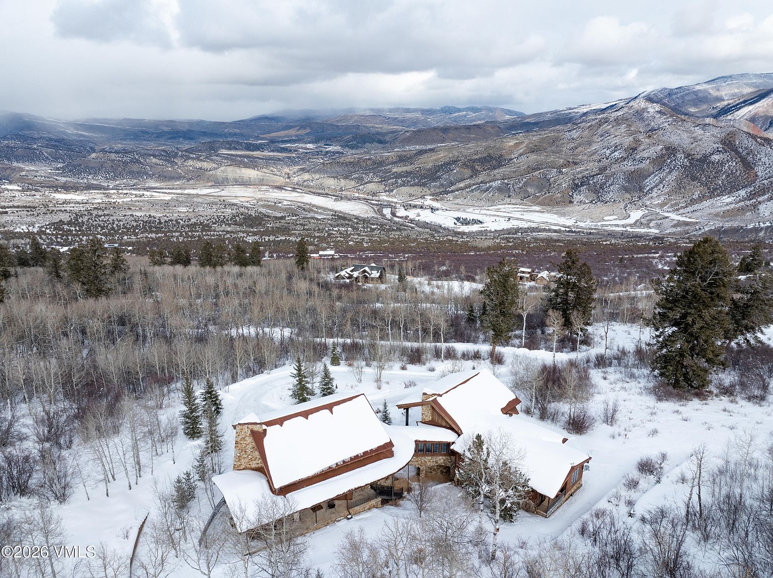 This aerial view showcases a luxurious mountain home nestled in a snowy landscape. The house features a stone and wood exterior with snow-covered roofs, blending seamlessly with the surrounding trees and mountains in the background. The perspective gives a sense of seclusion and grandeur, highlighting the property's expansive setting.