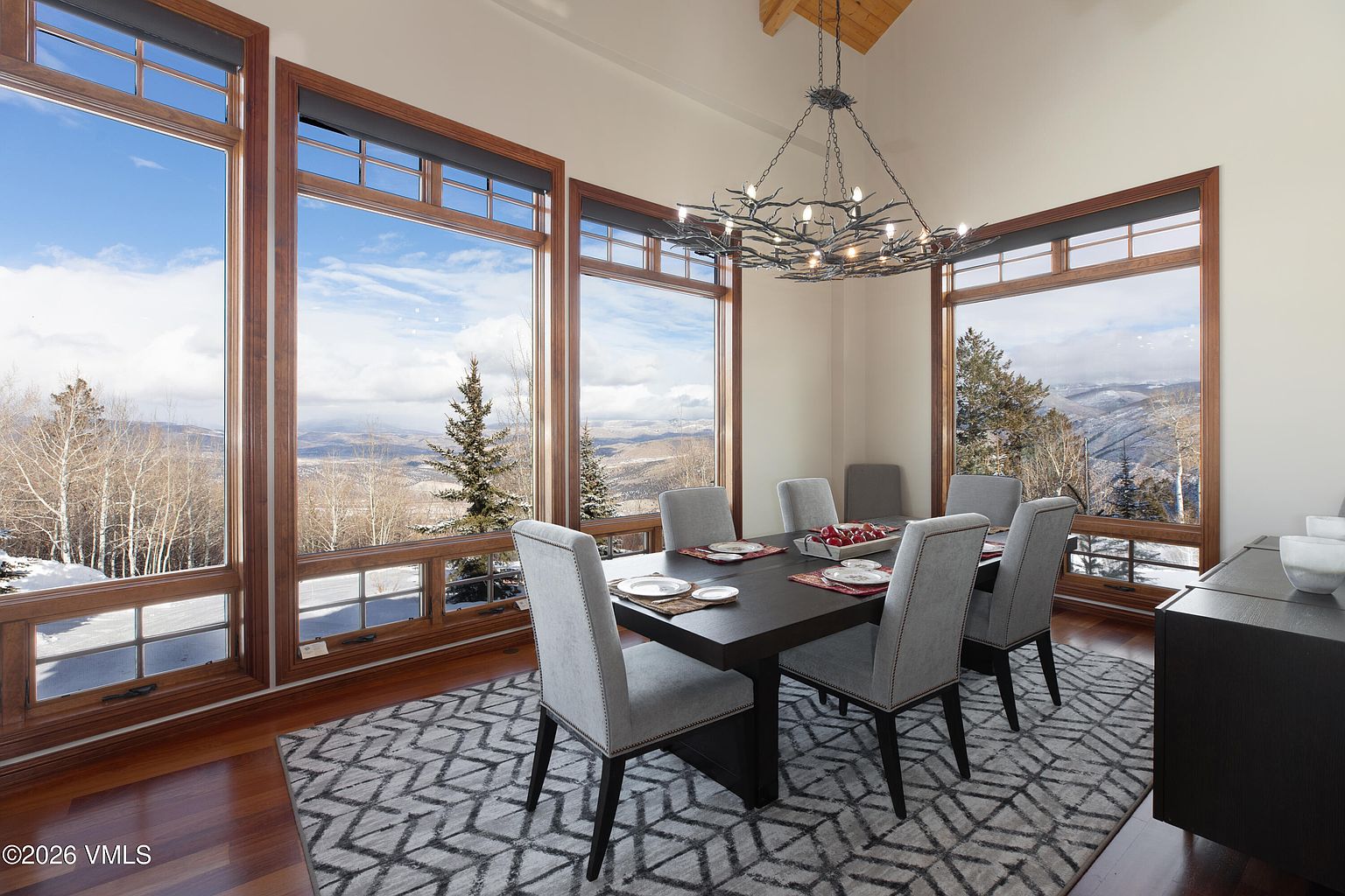 This is an interior shot of a dining room featuring a dark wood dining table with six upholstered chairs. Large windows offer a scenic view of a snow-covered landscape. An ornate chandelier hangs above the table, and a patterned rug covers the hardwood floor, adding texture and visual interest to the space.