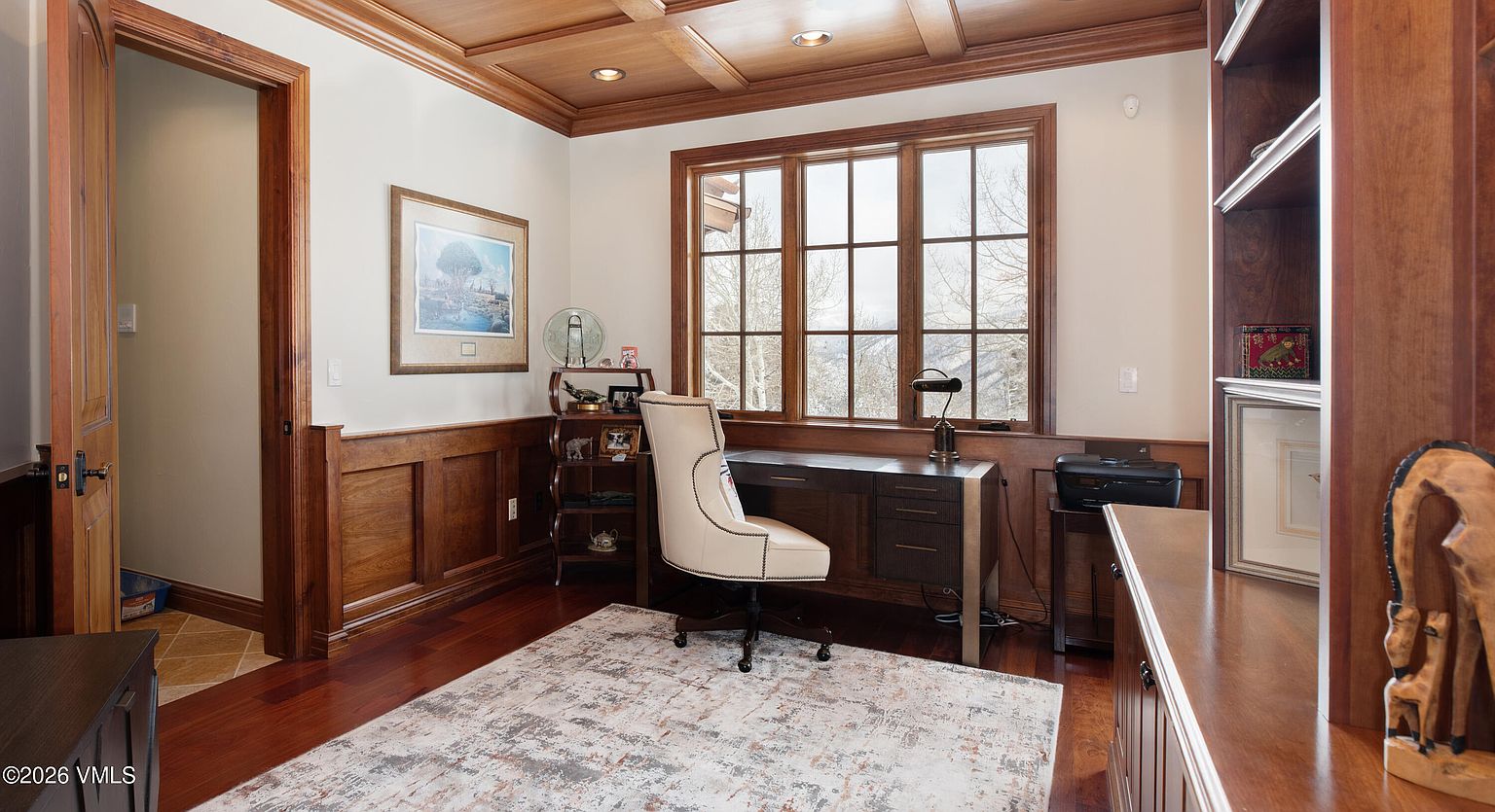 This is an interior shot of a home office or study. The room features wood paneling, a coffered ceiling, and a large window providing natural light. A desk and chair are centrally located, with a bookshelf visible on the right. The overall impression is one of a sophisticated and comfortable workspace.