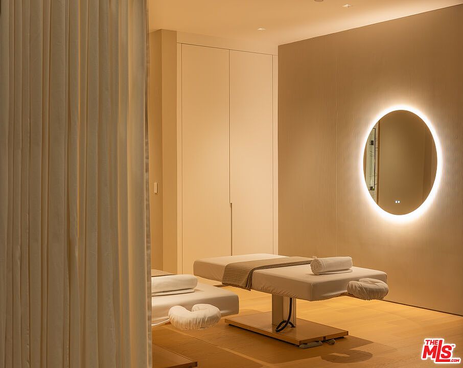 The image showcases a serene spa or massage room, featuring two massage tables with plush white linens and headrests. A large, circular mirror with integrated lighting adds a modern touch to the neutral-toned walls. The room exudes a sense of calm and relaxation, enhanced by the soft lighting and minimalist decor.