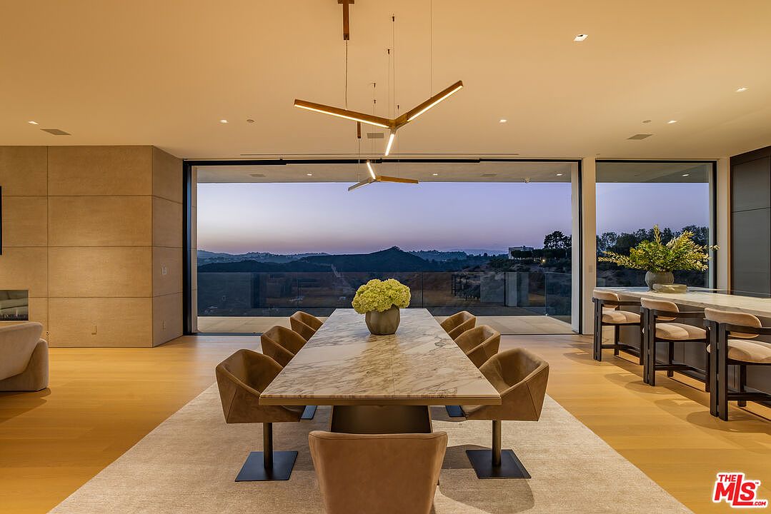 This is an interior shot of a modern dining room featuring a long marble-top dining table with seating for ten. A unique, multi-tiered chandelier hangs above the table, and a large sliding glass door provides a panoramic view of the landscape. The room is spacious and well-lit, creating an elegant and inviting atmosphere.