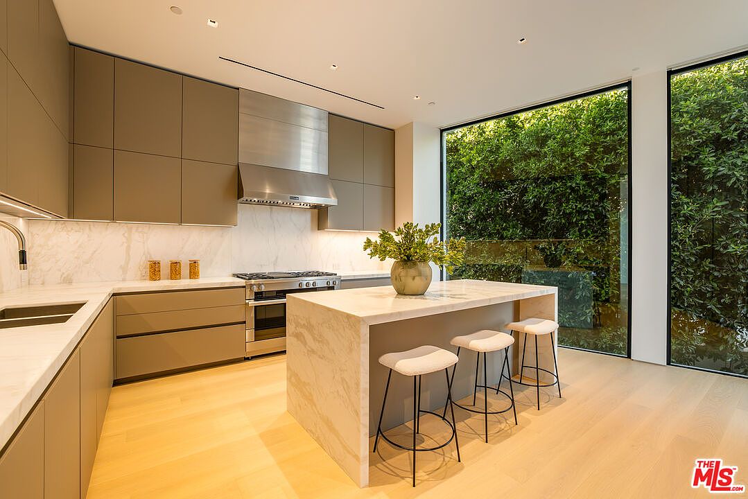 This is a modern kitchen with sleek, handleless cabinetry in a neutral tone, complemented by marble countertops and backsplash. A stainless steel range hood is positioned above the stove. The kitchen island features a marble waterfall countertop and three bar stools, while large windows offer a view of lush greenery, creating a bright and airy atmosphere.