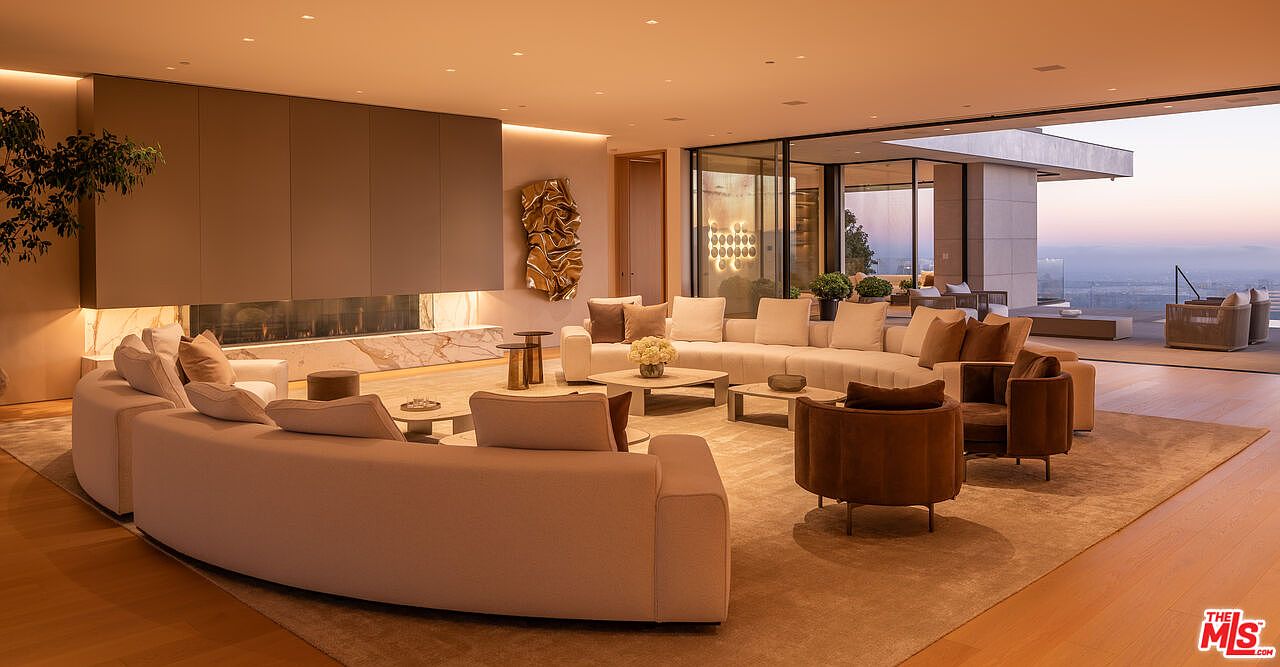 This is a luxurious living room featuring a curved sofa, modern art piece, and a fireplace. The room has an open floor plan with large glass doors leading to an outdoor patio area, offering stunning views. The color palette is neutral, creating a warm and inviting atmosphere.