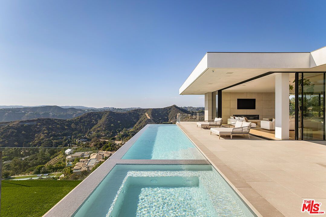 This image showcases a luxurious infinity pool and spa area of a modern home, offering stunning panoramic views of the surrounding mountains and landscape. The pool seamlessly blends with the horizon, creating a sense of serenity and sophistication, while the adjacent patio features stylish outdoor furniture, perfect for relaxation and entertainment. The architecture is sleek and contemporary, emphasizing clean lines and open spaces.