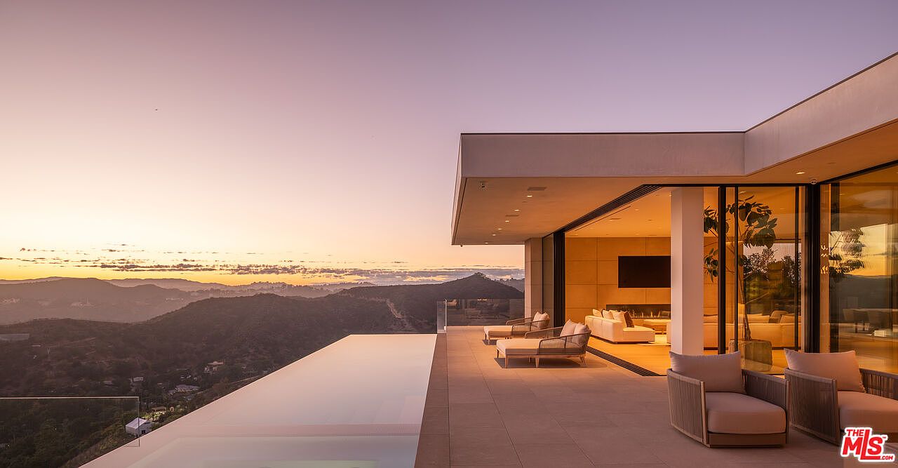 This image showcases a luxurious patio or balcony area with stunning views of the mountains and sunset. The space features modern outdoor furniture, including lounge chairs and armchairs, arranged on a tiled surface. An infinity pool adds to the elegance, while large glass doors provide seamless access to the interior, creating an inviting indoor-outdoor living experience.