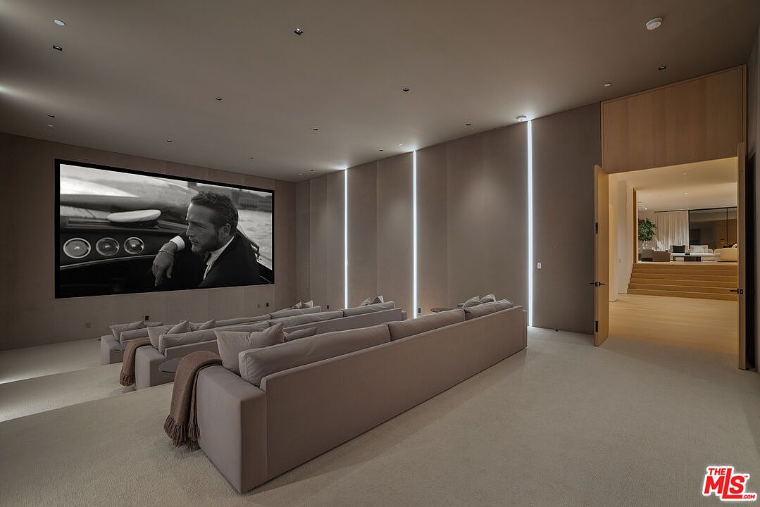 This is a luxurious home theater featuring tiered seating with plush, neutral-toned sofas and a large screen displaying a black and white film. The room is illuminated by vertical light strips integrated into the walls, creating a modern and sophisticated ambiance. An open doorway leads to another living space, suggesting a seamless flow within the home.