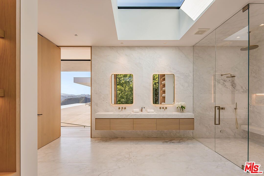 This is a luxurious primary bathroom featuring a double vanity with a marble countertop and wooden cabinets. Two rectangular mirrors with gold frames hang above the vanity, reflecting natural light from a skylight. A glass-enclosed shower with marble walls and a built-in bench is visible to the right, creating a spa-like atmosphere.