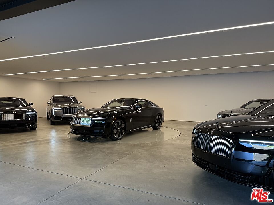 The image showcases a spacious garage featuring several luxury Rolls Royce vehicles. The garage has a minimalist design with a light gray concrete floor and white walls, illuminated by modern strip lighting on the ceiling. The overall impression is one of luxury and sophistication, highlighting the ample space and high-end aesthetic.