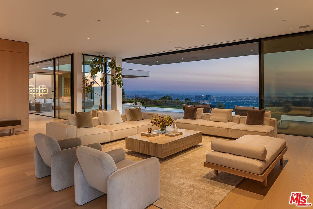 This is a luxurious living room featuring a large sectional sofa, modern armchairs, and a stylish coffee table. Floor-to-ceiling windows offer panoramic views of the city skyline, creating a seamless indoor-outdoor living experience. The room is bathed in natural light, highlighting the neutral color palette and contemporary design.