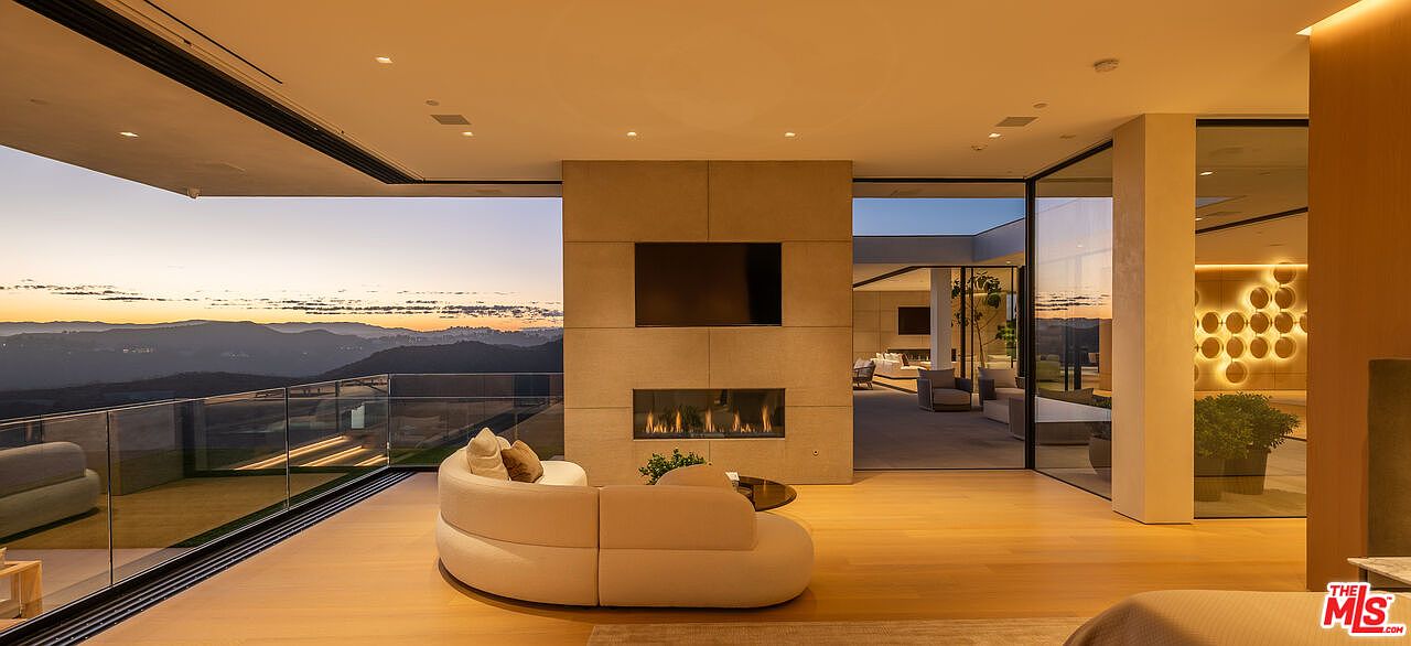 This is a luxurious living room featuring a modern fireplace with a TV above it, a curved sofa, and floor-to-ceiling windows offering panoramic views of the mountains at sunset. The open floor plan seamlessly connects the interior with the outdoor balcony, creating a sense of spaciousness and sophistication. The warm lighting and hardwood floors add to the inviting atmosphere.