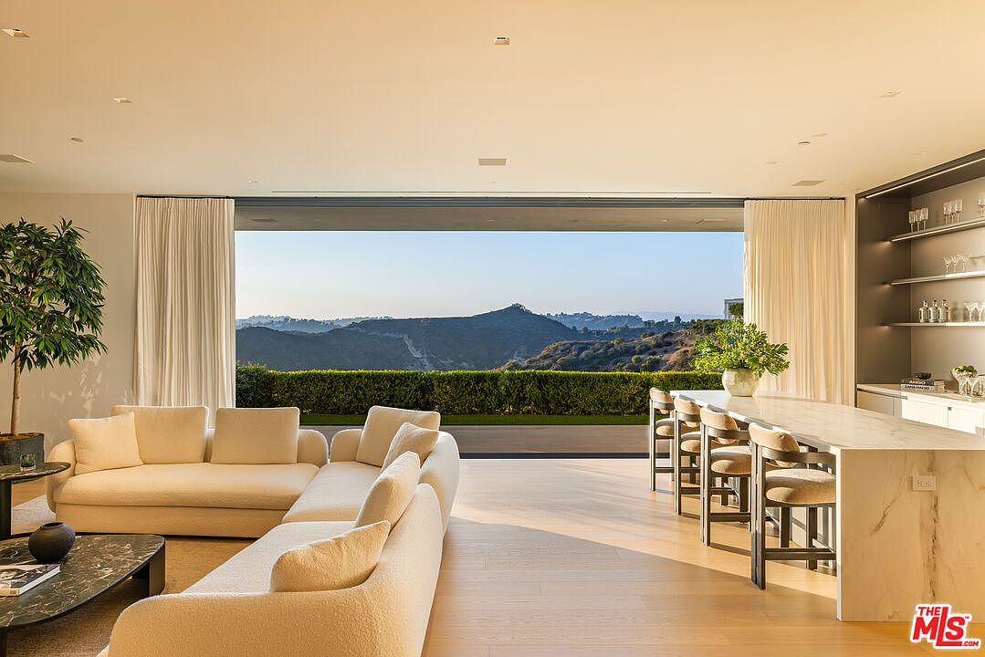 This image showcases a luxurious living room with an open floor plan, seamlessly blending indoor and outdoor living. A large, comfortable sectional sofa anchors the space, while floor-to-ceiling windows offer breathtaking views of the surrounding landscape. A modern kitchen island with bar seating is adjacent to the living area, creating an inviting and sophisticated atmosphere.