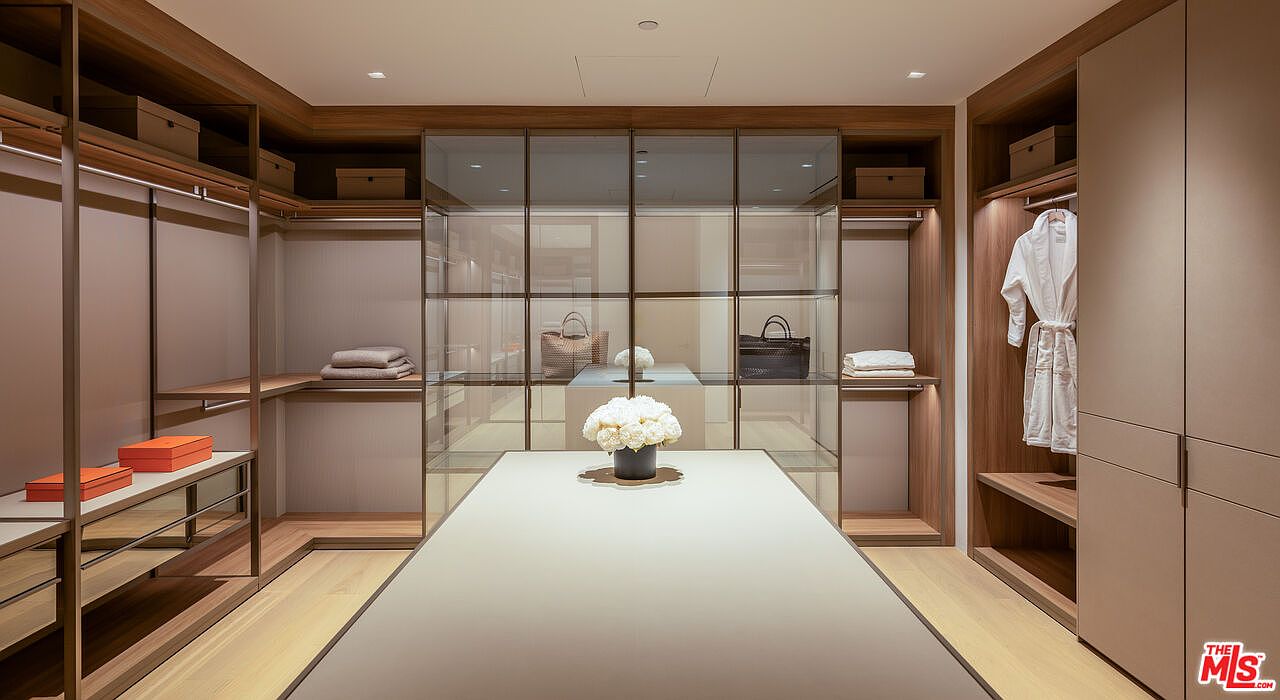 This is a well-organized walk-in closet featuring custom shelving, hanging rods, and a central island with a light-colored countertop. The closet is illuminated by recessed lighting, and the overall design is modern and luxurious. The closet includes glass-fronted display cases and a variety of storage solutions.