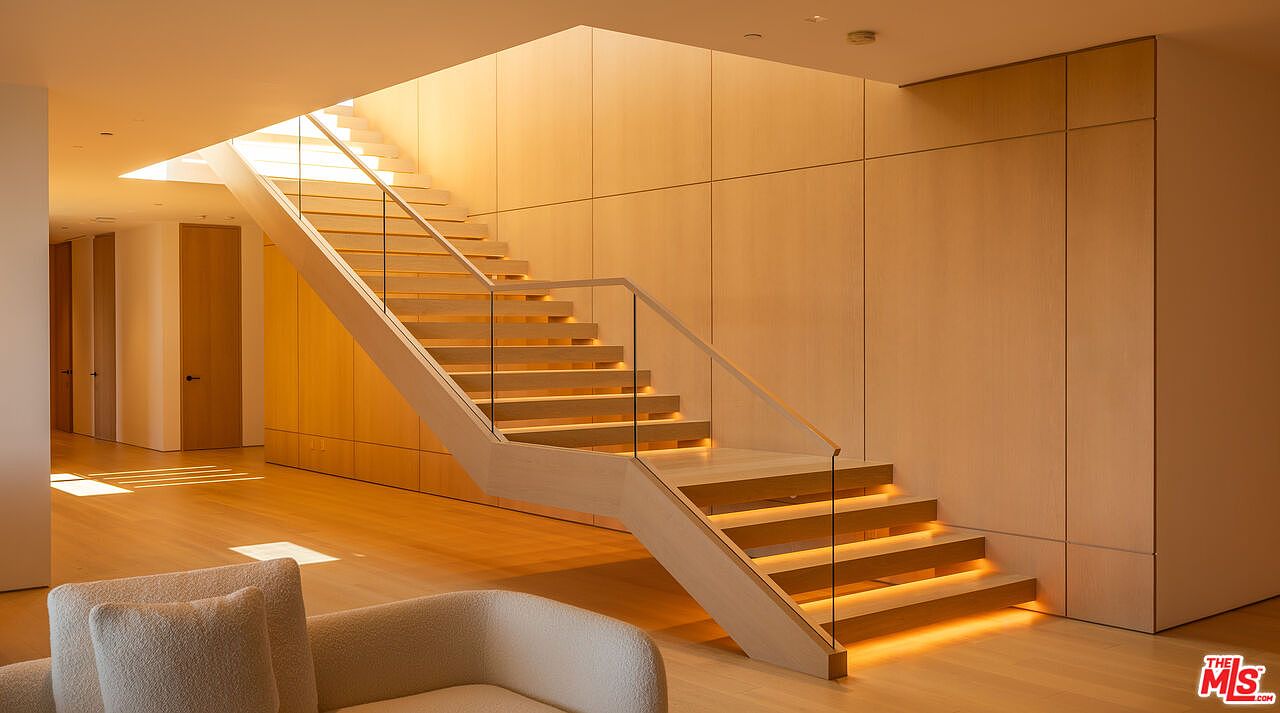 This interior shot showcases a modern staircase with wooden steps and glass railings, illuminated by natural light from above. The surrounding walls are clad in light wood panels, creating a warm and inviting atmosphere. The design emphasizes clean lines and minimalist aesthetics, perfect for a contemporary home.