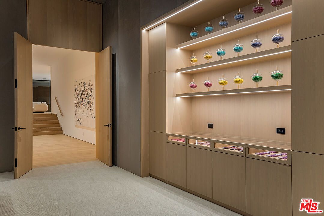 This interior shot showcases a modern hallway with a unique built-in display cabinet featuring illuminated shelves and glass-topped storage. An open doorway leads to a staircase and a glimpse of a living area, creating a sense of depth and flow. The neutral color palette and clean lines contribute to a sophisticated and minimalist aesthetic.